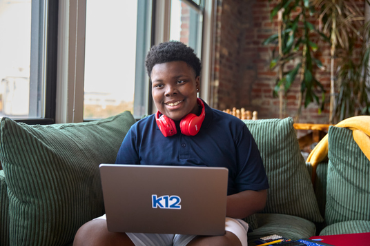 Person sitting on a couch with a laptop featuring the K12 logo, wearing red headphones.